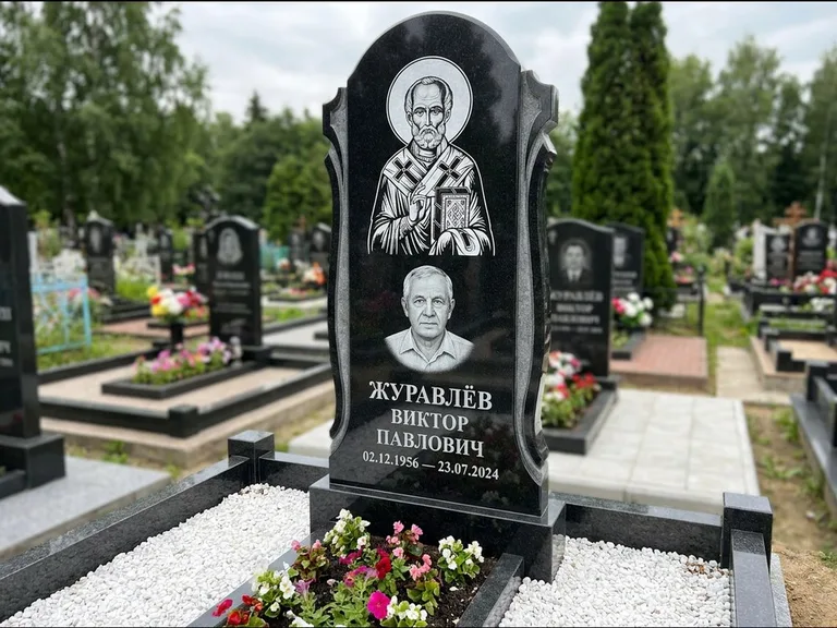 Памятник с гравировкой иконы: стела с образом в полный рост на обороте — гравировка образа Матроны Московской, низкие кусты спиреи в цвету, работа «Лебедева» в Магнитогорске
